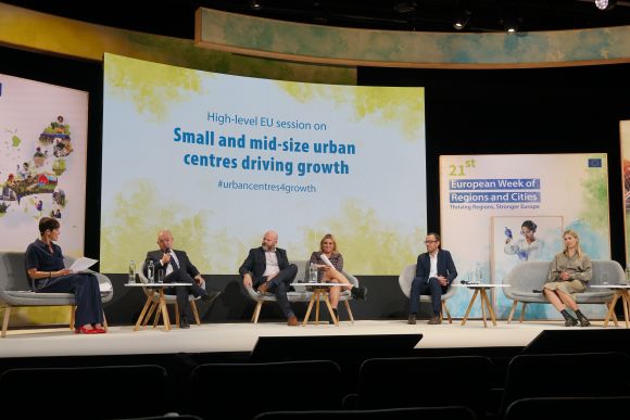 Speakers on stage in front of a screen reading "High-level EU session on Small and mid-size urban centres driving growth"